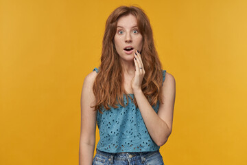 Indoor portrait of young redhead female with wavy long hair touching her cheek looks into camera with shocked, surprised facial expression, isolated over orange background