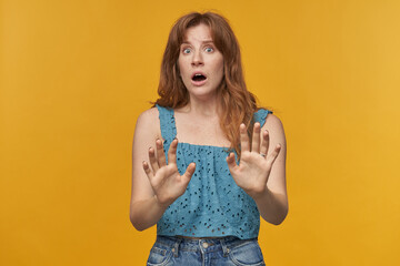 Indoor portrait of young redhead female with wavy long hair shows stop negative sing with her hands with irritated facial expression isolated over orange background