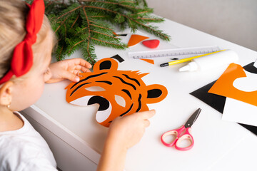 a little girl makes a New Year's tiger mask from colored paper, the symbol of 2022, with her own hands