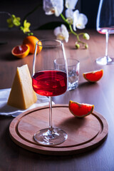 Elegant goblet with rose wine, cheese and oranges on wooden surface in romantic setting against backdrop of blooming orchids.