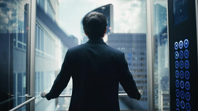 Stylish Japanese Businessman Riding Glass Elevator to Office in Modern Business Center. Male Looking at Modern Downtown Skyscrapers Out of the Panorama Window in the Lift. Back Turned to Camera.