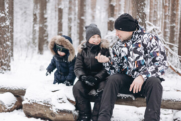 Fototapeta na wymiar Happy father and little sons enjoying talking sitting together on log in winter snowy forest. Joyful man and teenager boy resting bonding having friendly positive conversation on frosty day outdoors