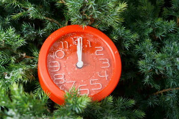 Red clock with white hands at 12 o'clock and 1 second with snowflakes on glass among green pine...