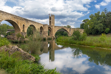 Fototapeta na wymiar Besalu, Spain, medieval stone bridge with ancient gate, dramatic sky with clouds, mrsta reflection in the water