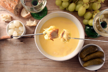 Melted cheese fondue with bread on long forks, pickles, grapes and white wine on a rustic wooden table, copy space, high angle view from above, selected focus