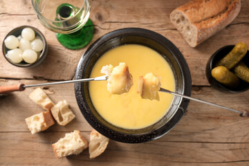 Fondue from melted cheese with bread on long forks, pickles and wine on a rustic wooden table,...