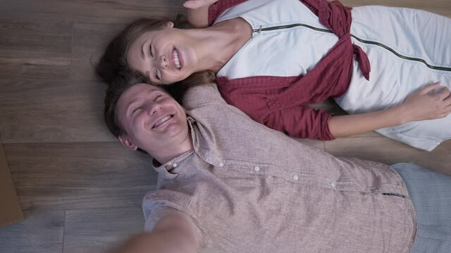 Selfie camera POV cheerful couple smiling taking photo lying on floor indoors. Cheerful beautiful woman and joyful man having fun photographing on parquet in slow motion waving looking at camera.