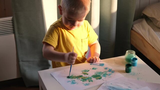 Little Boy Draws Picture On Paper. Preschool Child Drawing with Paints at the Table at Home. Green Paint, Yellow Clothes. Day Sunlight. Real People. Creative and Artistic little Kid.	
