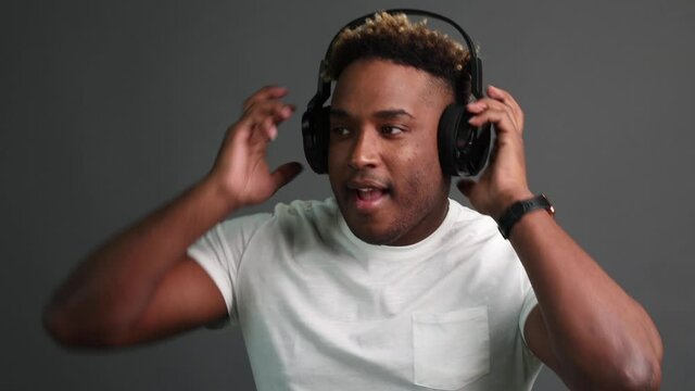 African guy in a white T-shirt listens to music with black headphones and dances on a gray studio background. Charismatic man with an African hairstyle lights up in the dance
