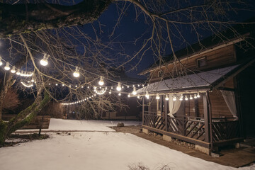 Wooden country house with a veranda on the plot in winter. Outside view. Lighting the site with a garland of light bulbs.