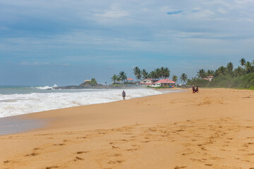 Fototapeta na wymiar Asia. Sri Lanka. Ahungalla Beach. People walk along the sea with waves. Yellow sand. Blue sky. Beach houses with tiled roofs and palm trees.