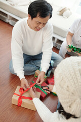 Smiling mature man helping wife to wrap Christmas presents and decorate with ribbon