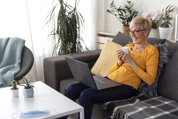 An older woman with short blonde hair sits on sofa in living room and chats with friend on laptop over video chat