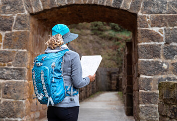 A girl is doing archeological tourism at an archaeological site. She is wearing a hat and a blue backpack and is looking at a map.