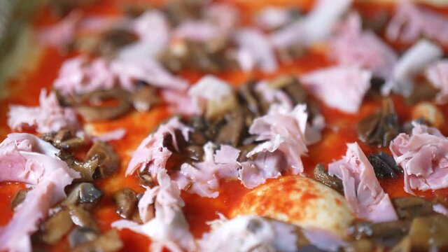 Closeup dolly shot of an organic traditional Italian pizza seasoned topped with ham and mushrooms on the dough with red tomato sauce on it.