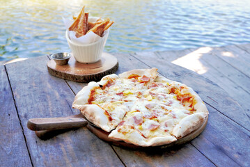 Hawaiian pizza with fried spring rolls on wooden deck beside lake