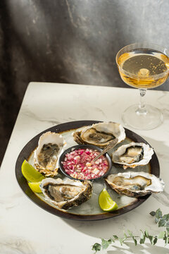 Fresh oysters with lime, onion on a grey round plate. Oyster season. Macro seafood dish. Oysters on the half shell with glass of sparkling wine on white table, beautiful light. Healthy food concept.