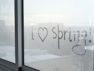 The misted window pane has the inscription "I love spring". The outlines of houses, roofs, snow can be seen through the glass. It is getting dark.