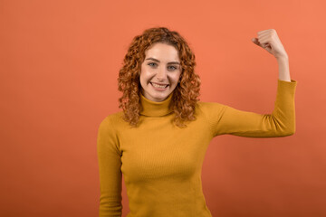 Young and attractive redhead Caucasian girl in orange jumper showing biceps isolated on orange studio background.