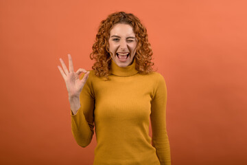 Young and attractive redhead Caucasian girl in orange jumper showing fingers Okay gesture isolated on studio orange background.