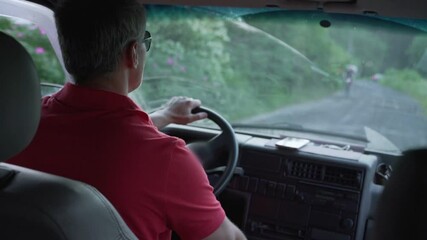 man with gray hair in sunglasses drives along dirt road in minivan during summer day