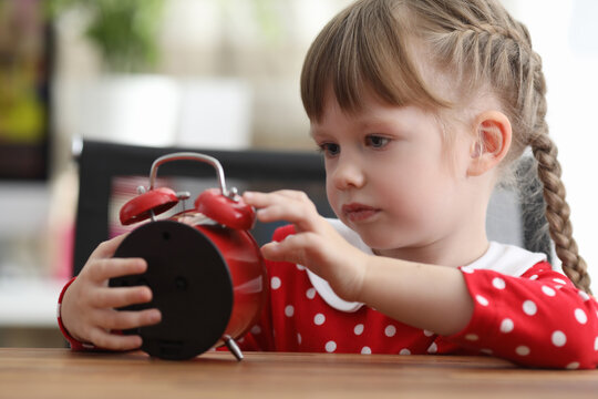 Little girl with pigtails looking at red alarm clock at table