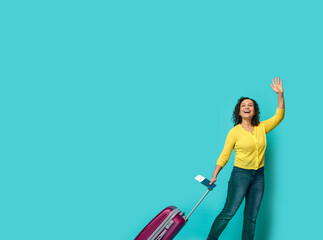 Full length of beautiful woman in casual clothes, waving hand, holding identity document and boarding pass, posing with suitcase, smiling looking to the side at a blue background with copy space