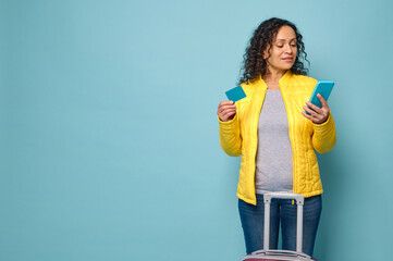 Young woman in yellow jacket stands against blue wall background, in front of her suitcase, holds mobile phone and plastic card with blank space for ad. Travel, booking, banking concept