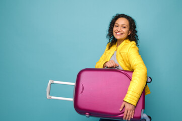 Charming African American woman traveler in bright yellow jacket, smiling with cheerful toothy smile looking at camera and holding her suitcase under arms, isolated on blue background with copy space