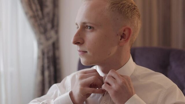 Businessman buttoning shirt near window in the morning. Stylish man groom straightens his white shirt preparing to go out. Guy get ready before date or meeting with bride fashion style weight fit