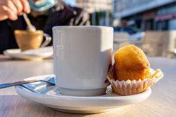 Close-up of a white cup of coffee, next to a small muffin, on a coffee shop table.