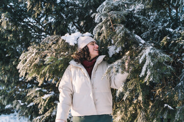 Young curly smiling woman walk in winter park and hide behind snow-covered Christmas tree. Positive emotions, freshness, holidays