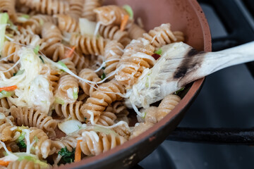 closeup of whole wheat pasta, vegetables, and melted cheese cooking in a orange ceramic pan on a stove and being stirred by a wooden cooking spoon