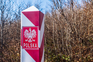 Polish, white and red border post. On the pole, there is a white eagle with a golden crown and the inscription Polska - Poland. In the background, the blue sky and trees, bushes and grass. - 477203370
