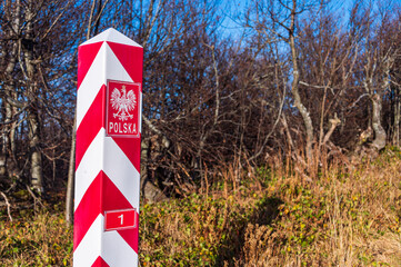 Polish, white and red border post. On the pole, there is a white eagle with a golden crown and the inscription Poland and number 1. In the background, the blue sky and trees, bushes and grass.