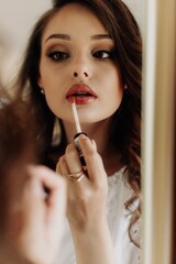 Reflection in the mirror of a beautiful brunette in a white dress with makeup who paints her lips with lip gloss..