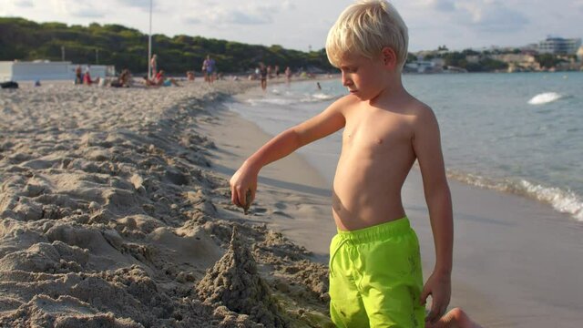 boy builds figure from wet sand on beach sea. Fokus of attention falling grains sand. The movement of water with a wave. Relaxation in independent play during the holidays. White hair.