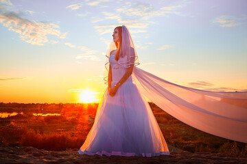 Beautiful bride in white dress and long fabric in the desert on sand on dunes and blue sky with yellow and red sunset