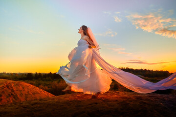 Fototapeta na wymiar Beautiful bride in white dress and long fabric in the desert on sand on dunes and blue sky with yellow and red sunset