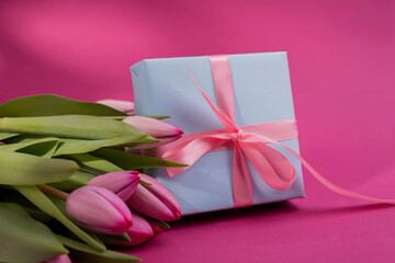 Romantic gift box tied with big pink bow and spring tulips on pink color background. Mother's day or Easter holiday concept.