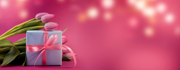 Romantic gift box tied with big pink bow and spring tulips on pink color background. Mother's day or Easter holiday concept.