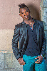 Dressing in a black blazer, green pants, a young black guy with mohawk hair is standing against the wall, his head tilted one side, confidently looking at you..