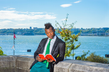 Man Reading Outside. Wearing a black blazer, green pants, a green neck tie, hands holding a red...