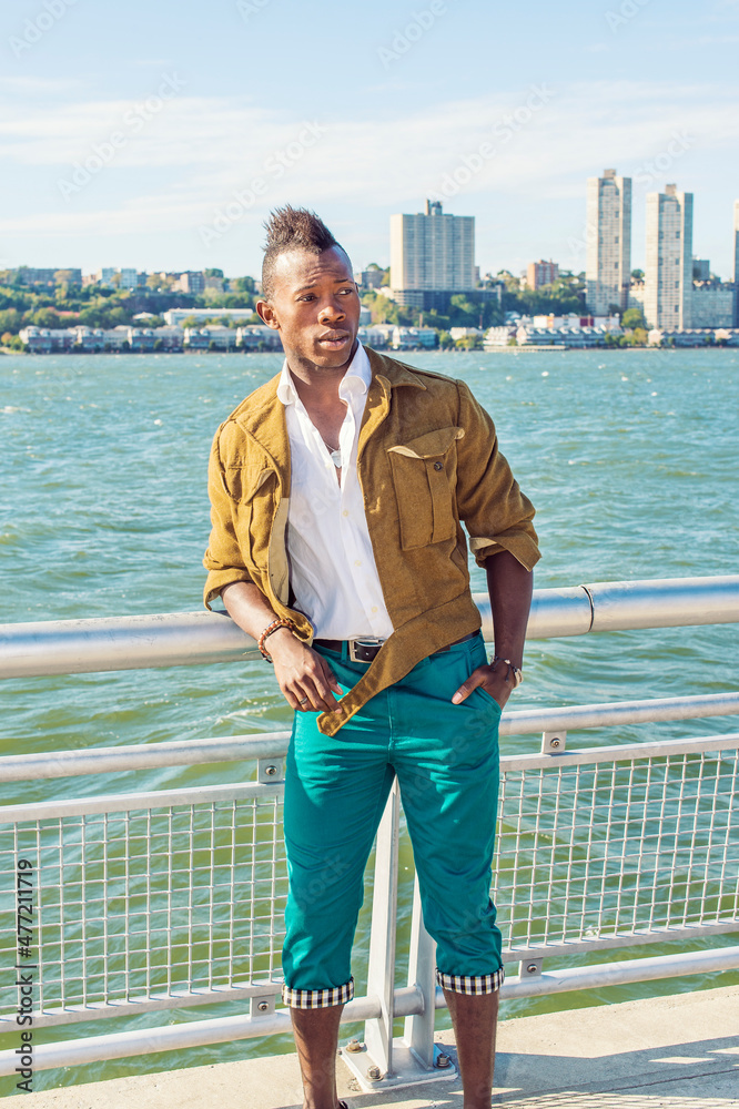 Wall mural Man Relaxing Outside. Dressing in a yellow woolen jacket, white underwear,  green pants, hands in pockets,  a young black guy with mohawk hair is standing by a river, looking around..