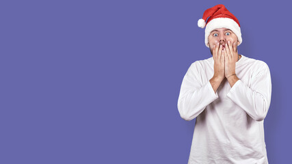 Fototapeta na wymiar a young man in a Santa Claus hat covers his mouth with his hands in shock