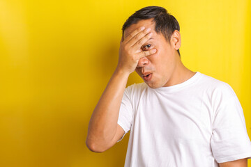 Asian man peeking between his fingers on yellow background 