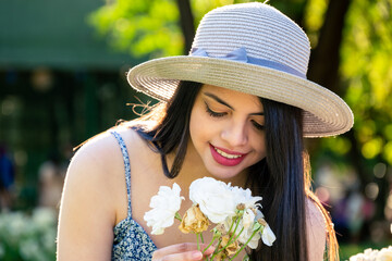Young woman at park using a hat smelling a flower during summer spring season at a sunny day. Brunette with long black hair.