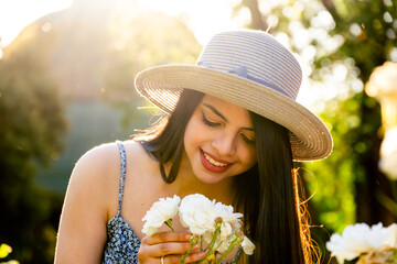 Young woman at park using a hat smelling a flower during summer spring season at a sunny day. Brunette with long black hair with a natural back light