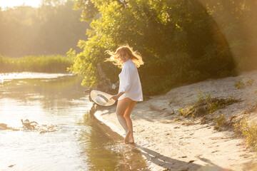 Portrait of young dreaming happy smiling woman weared white shirt and holding white hat walking near a lake or pond. Summer weekend or vacation story. Brown-haired girl with long hairs. Local tourism 