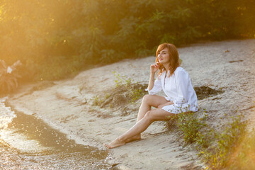 Portrait of young dreaming happy smiling woman weared white shirt and holding white hat walking near a lake or pond. Summer weekend or vacation story. Brown-haired girl with long hairs. Local tourism 